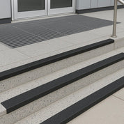 Modern commercial building entrance featuring terrazzo concrete steps with black 55mm anti-slip GRP nosings, stainless steel handrail, and metal drainage grate, providing durable slip resistance for public access.
