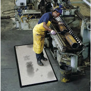 A worker in a yellow apron and blue shirt stands beside a large industrial machine, holding a tool near a metal roller. A rectangular gray mat with dark stains lies on the concrete floor in the foreground, and a bucket with a yellow bucket strap sits below.