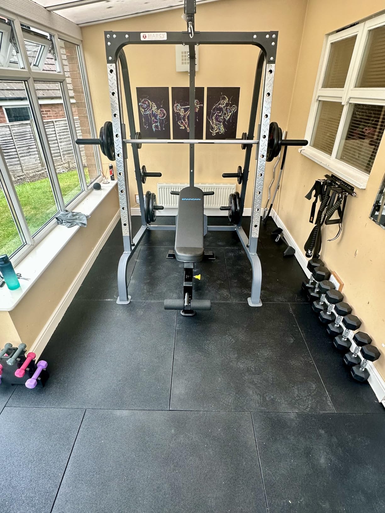 Home gym setup with weight rack, bench, and dumbbells in a room with large windows.
