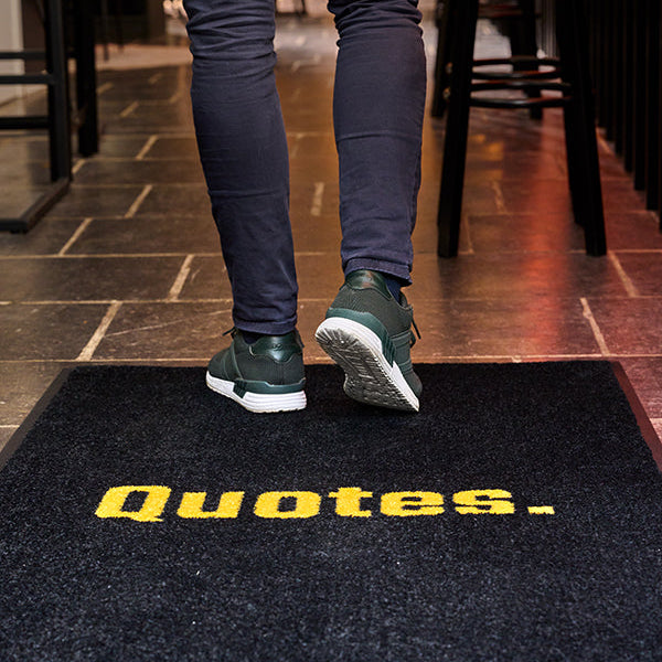 Person wearing dark pants and green shoes stepping onto a doormat with 'Quotes.' text.