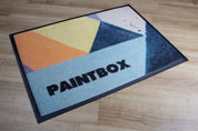 Geometric patterned mat with 'PAINTBOX' branding on a wooden floor.