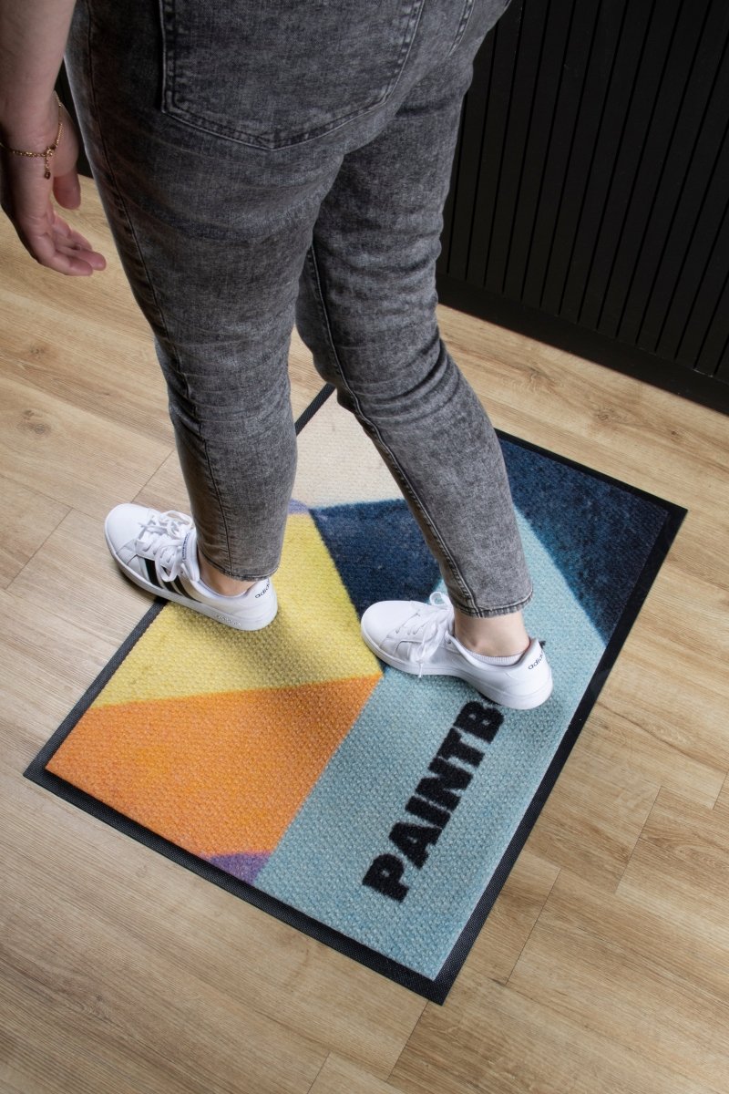 Person standing on a colorful doormat with 'PAINT' text on a wooden floor.