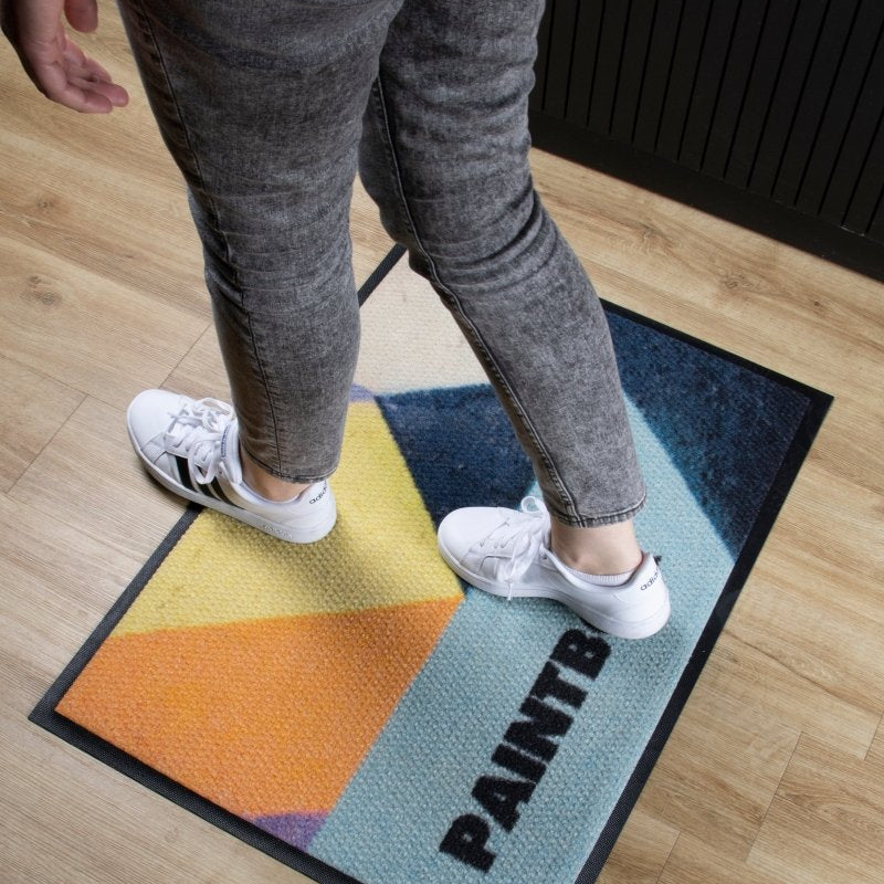 Person standing on a colorful doormat with 'PAINT' text on a wooden floor.