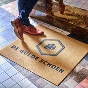 Person stepping onto a doormat with 'De Gulde Schoen' branding on a tiled floor.