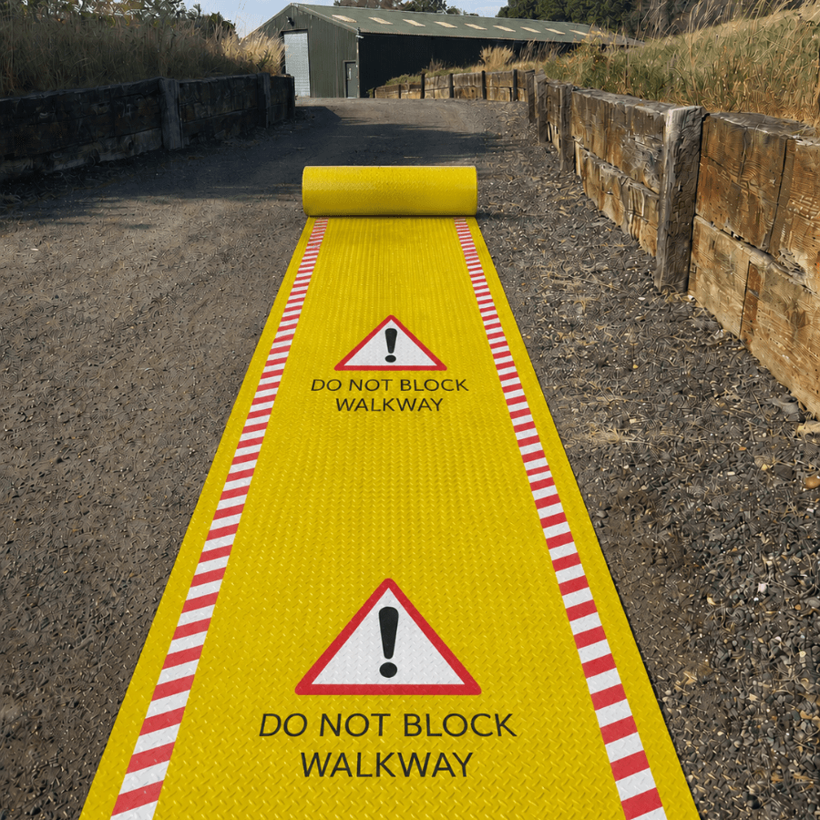 A bright yellow roll-up do not block walkway mat with red-and-white striped side borders and warning triangle signs, shown unrolled along a gravel path outdoors.