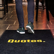 Person wearing dark pants and green shoes stepping onto a doormat with 'Quotes.' text.