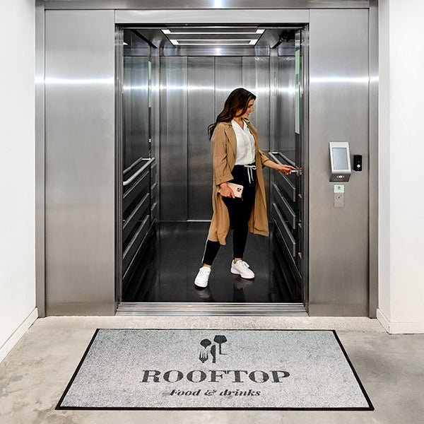 Woman entering an elevator with a 'Rooftop Food & Drinks' mat in the foreground.