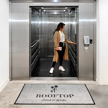 Woman entering an elevator with a 'Rooftop Food & Drinks' mat in the foreground.