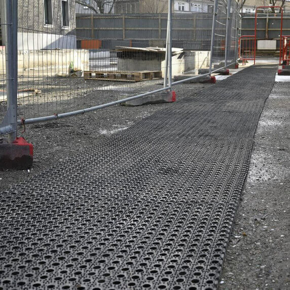 A black plastic grid drainage mat laid out on a construction site, with chain-link fencing and building materials visible in the background.