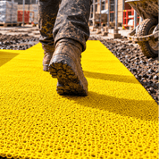 A construction worker in a high-visibility vest walks over a bright yellow textured mat laid out on a muddy construction site, with a rolled-up section and construction equipment in the background.