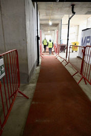 A red carpet runner guides down a concrete construction corridor, flanked by red metal crowd control barriers. Two construction workers in high-visibility yellow vests are visible in the distance.