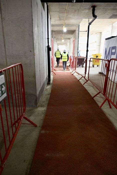 A red carpet runner guides down a concrete construction corridor, flanked by red metal crowd control barriers. Two construction workers in high-visibility yellow vests are visible in the distance.