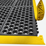 Close-up of a black rubber anti-slip grid mat with a bright yellow metal edge and corner protectors, shown on a light gray background.