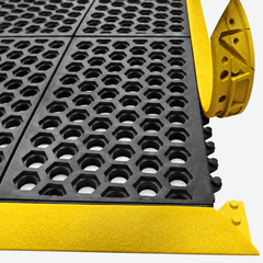 Close-up of a black rubber anti-slip grid mat with a bright yellow metal edge and corner protectors, shown on a light gray background.