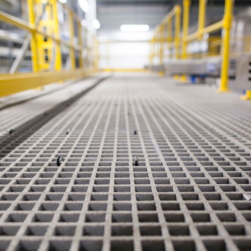 Close-up of a GRP MESH grid-like surface in an industrial setting with blurred yellow metal structures in the background.