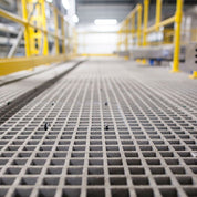 Close-up of a GRP MESH grid-like surface in an industrial setting with blurred yellow metal structures in the background.