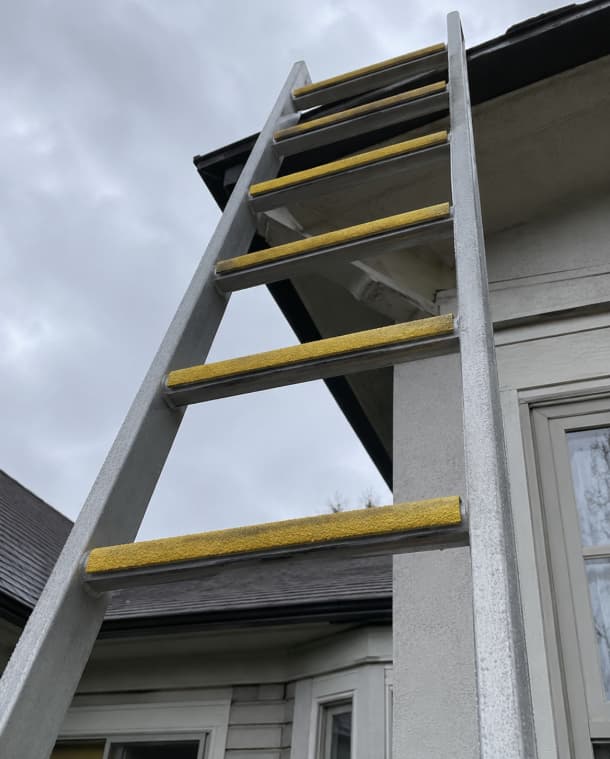 A gray metal ladder with textured yellow hand grips positioned against the exterior wall of a house under an overcast sky.